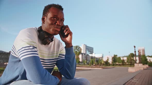 A Young Black Student in Stylish Clothes Sits and Talks on a Smartphone in the City