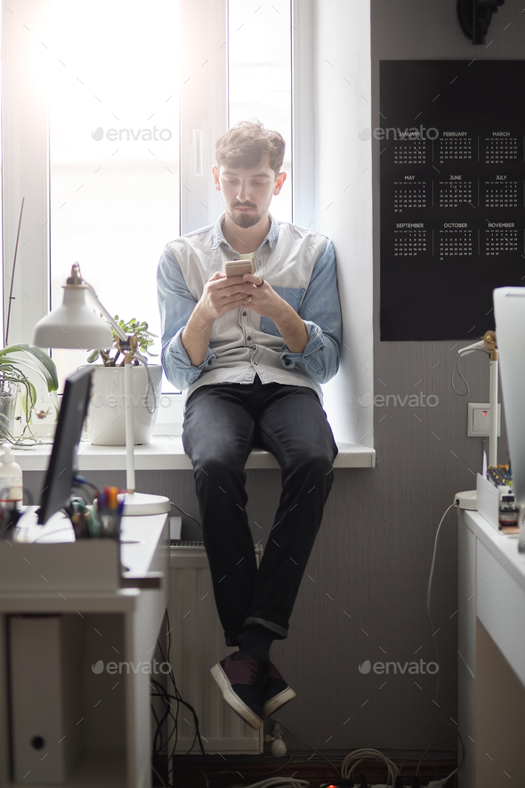 Young stylish man sitting on the window with phone in hands Stock Photo by arthurhidden
