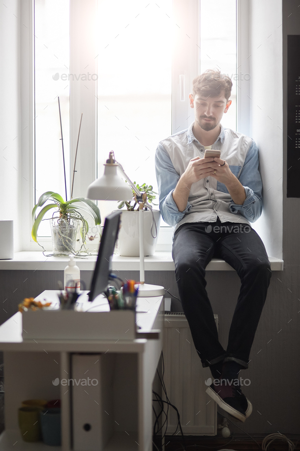 Young stylish man sitting on the window with phone in hands Stock Photo by arthurhidden