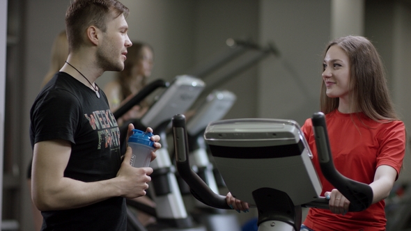 Girl in Red Shirt Vigorously Works on Exercise Bike and Guy Comes To Ask Her Name