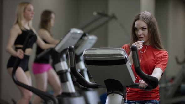 Awesome Girl in Red Shirt Vigorously Works on Exercise Bike and Talks with Her Phone with Headphone