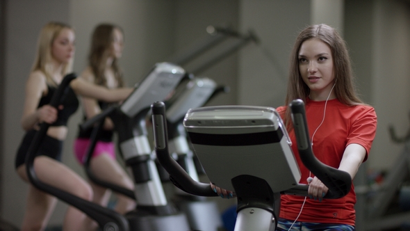 Lovely Girl in Red Shirt Vigorously Works on Exercise Bike and Talks with Her Phone with Headphone