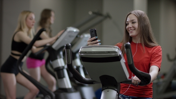 Nice Girl in Red Shirt Vigorously Works on Exercise Bike and Talks with Her Phone in the New Gym