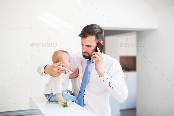 Father with shirt and tie and smartphone feeding a baby son at home, when talking on the phone. Stock Photo by halfpoint