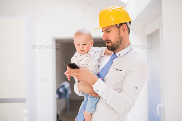 Father with shirt, tie, helmet and smartphone holding a baby son at home. Stock Photo by halfpoint