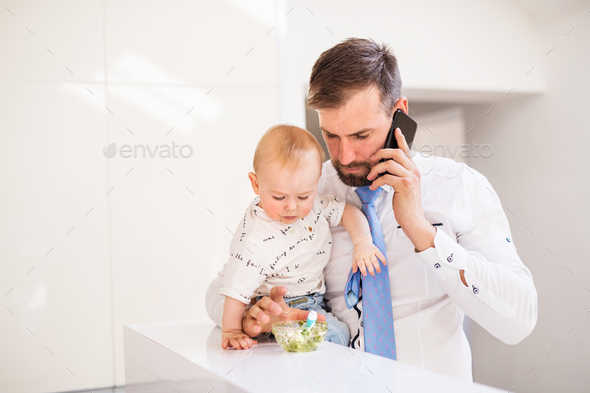Father with shirt and tie and smartphone feeding a baby son at home, when talking on the phone. Stock Photo by halfpoint