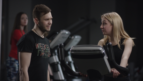 Awesome Girl in Blak Sport Wear Vigorously Works on Exercise Bike and Guy Comes To Show His Muscular