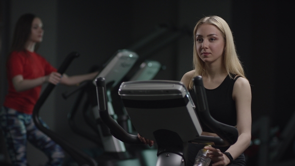 Awesome Girl in Blak Sport Wear Vigorously Works on Exercise Bike and Listens Music with Headphones