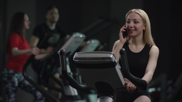 Nice Girl in Blak Sport Wear Vigorously Works on Exercise Bike and Talks with Her Phone in the New