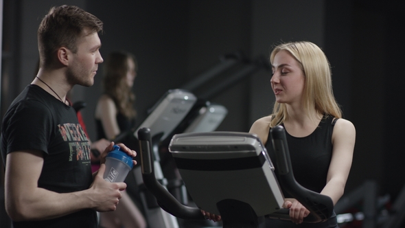 Nice Girl in Blak Sport Wear Vigorously Works on Exercise Bike and Guy Comes To Meet and To Ask Her