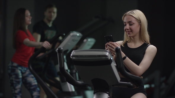 Nice Girl in Blak Sport Wear Vigorously Works on Exercise Bike and Texting Message in Chat with Her