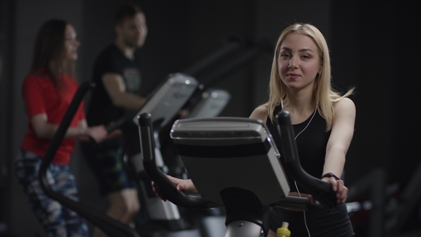 Cute Girl in Blak Sport Wear Vigorously Works on Exercise Bike and Approve Showing Thumbs-up in the