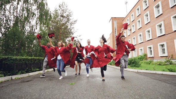 Dolly Shot of Joyful Graduating Students Running with Diplomas Waving Mortar-boards and Laughing alt