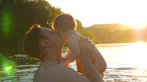 Happy Parents Bathe an Infant Who Smiles and Laughs in Splashes at Glare of Setting Sun alt