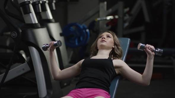 Nice Blonde Curly Girl with Straight Hair in a Black Sport Wear with Blue Eyes Does a Dumbbell Press