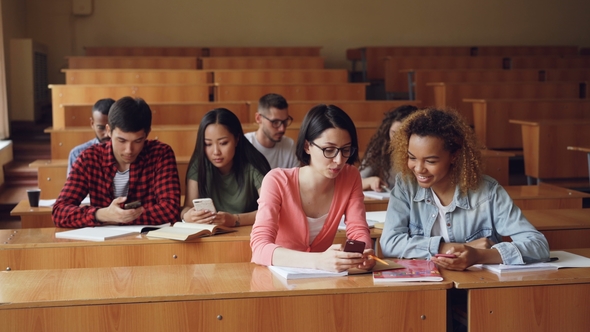 Young People Are Using Smartphones and Talking Sitting at Desks in Lecture Hall at University alt