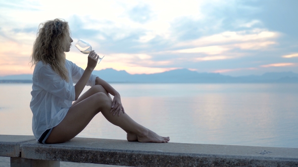 Young Woman Sitting on the Beach and Meets a Sunset with a Glass of Wine in Her Hand alt