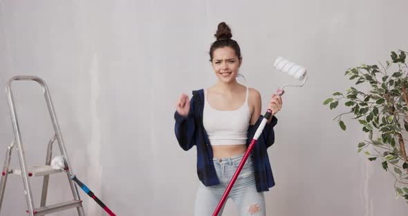 Smiling Pretty Girl Dressed in Tight Clothing Shirt Dances with Paint Roller Stick on Walls Beside alt