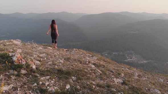 A Young Woman Stands on the Edge of a Rock and Raises Her Hands