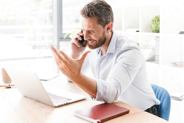 Happy handsome bearded man talking by mobile phone while using laptop computer. - Stock Photo - Images