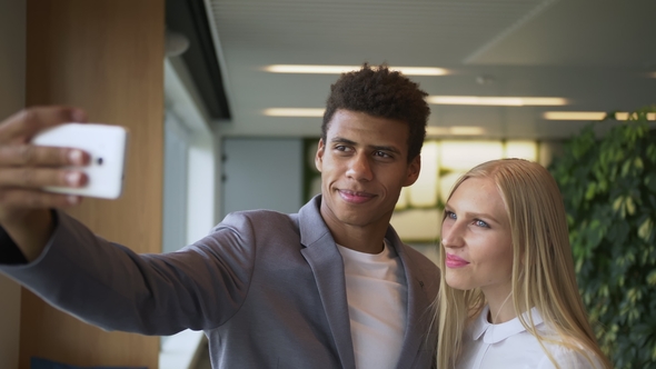 Laughing Diverse Coworkers Taking Selfie in Office. Cheerful Black Man with Laughing Blond Woman alt
