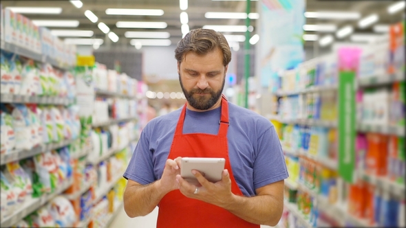 Handsome Salesman Using Digital Tablet Standing Among Shelves In Supermarket