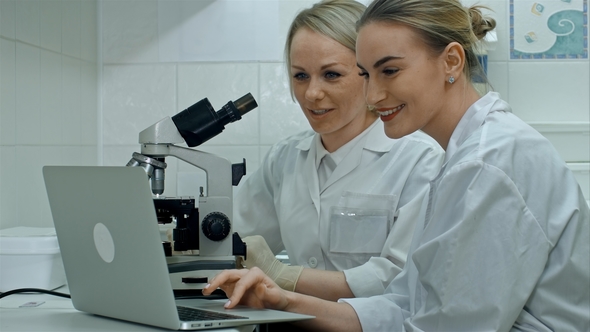 Two Young Positive Scientists Working in Laboratory Using Microscope and Laptop alt
