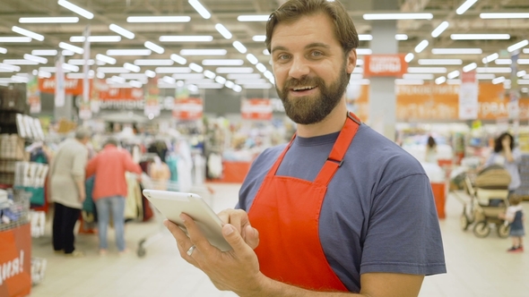 Handsome Supermarket Clerk Using a Touch Screen Tablet in Supermarket, He Is Smiling at Camera