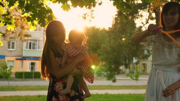 Mom and Her Daughters Catch Big and Transparent Soap Bubbles in City Park and Laugh in Glare of Sun