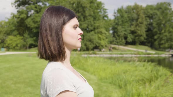 A Young Caucasian Woman Looks Around Thoughtfully By a Pond in a City Park on a Sunny Day alt
