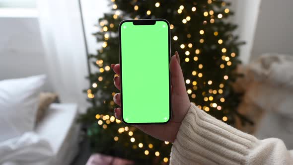 Woman holding modern smartphone with green screen chromakey near Christmas tree lights on background