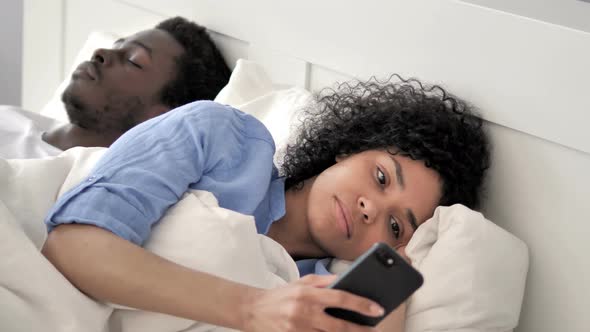 African Woman Using Smartphone While Boyfriend Is Sleeping alt