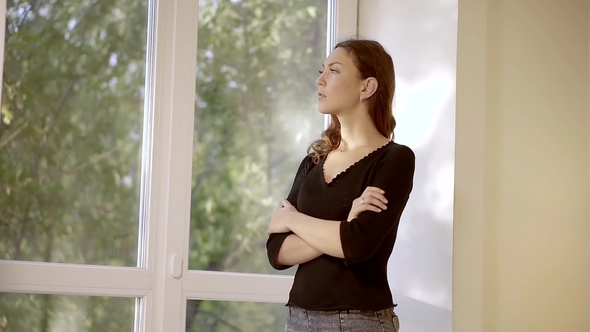Sad Young Girl Standing at the Window of His House with Folded Hands at the Breast alt