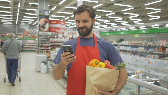 Smiling Supermarket Employee Using Mobile Phone and Holding Shopping Bag in Supermarket