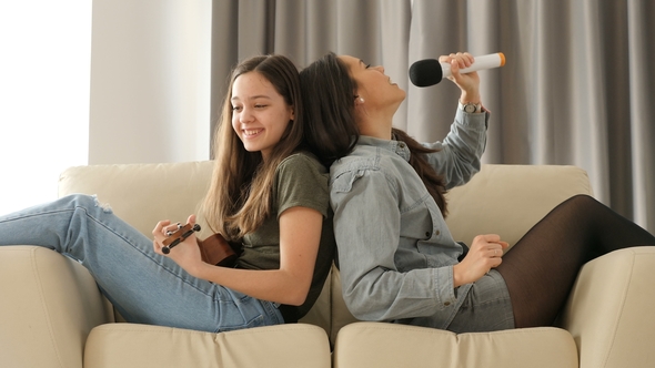 Two Sisters Having Fun on the Couch Playing at Ukulele and Singing alt