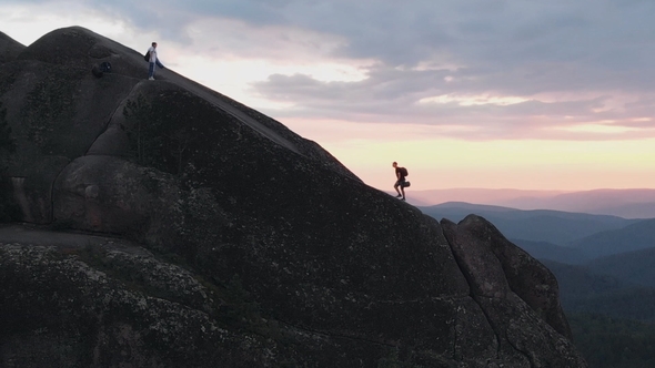 Aerial Shot of Two Friends of Tourists Climb a High Rock at Sunset.