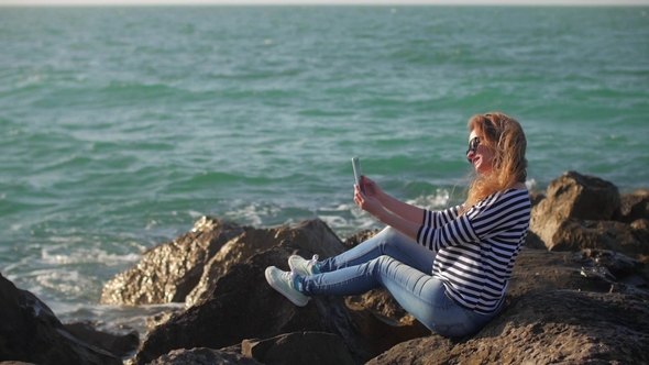 Blonde Girl with Tablet in Hands Is Sitting on a Huge Stones Shore in Sunny Summer Day