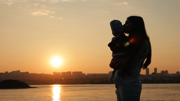 A Young Mother Kisses Her Little Son While Standing By the River at Sunset