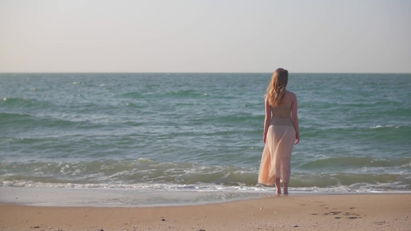 Slim Blonde Girl Is Standing on a Sand Beach in Seawater and Watching Waves in Summer Day