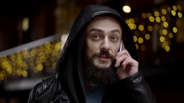 Bearded Man in a Leather Hooded Jacket Speaks Emotionally Over a Cell Phone alt
