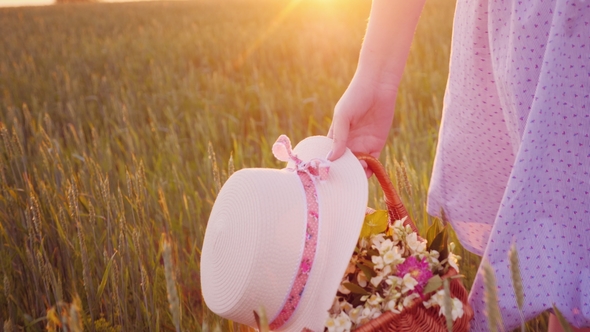 A Woman Carries a Basket in Field Flowers and a Hat Over a Green Field alt