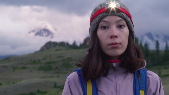 A Young Woman with a Backpack and a Headlamp Looks at the Camera Before Climbing. Foggy Mountains in