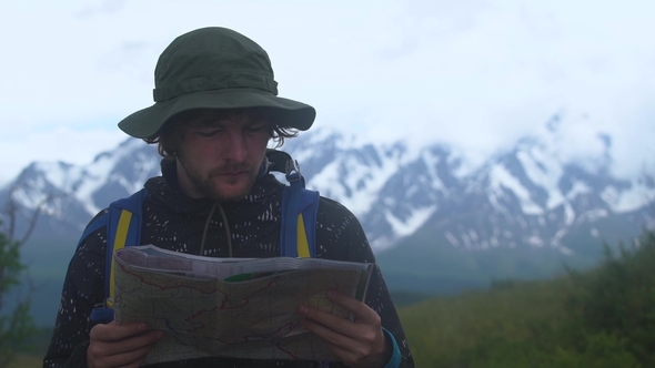 Sad Hipster Young Man with Backpack Holding in Hands and Looking on Map of Foggy Mountain, Tourist