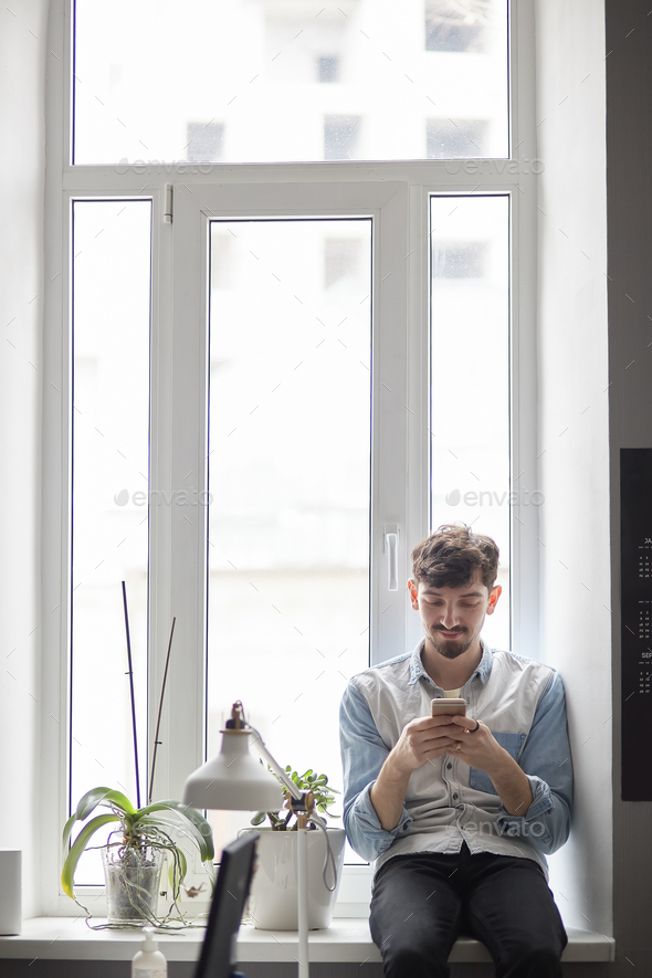 Young stylish man sitting on the window with phone in hands - Stock Photo - Images
