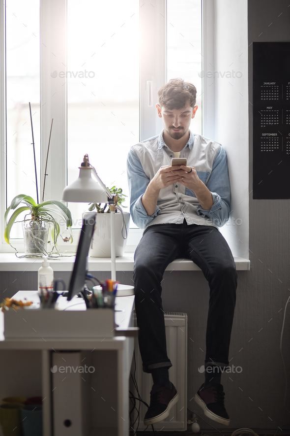 Young stylish man sitting on the window with phone in hands - Stock Photo - Images