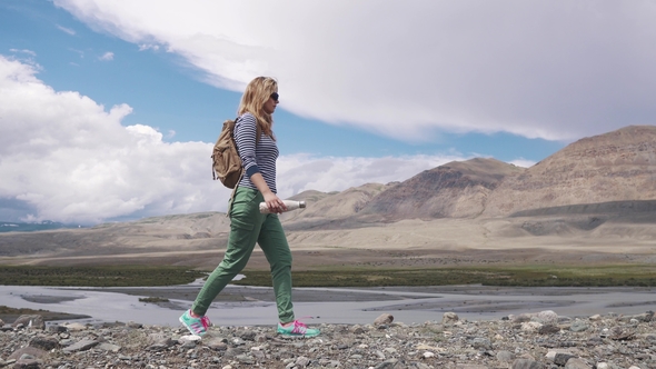 Traveler Walks Through the Mountainous Terrain. Girl Tourist with a Backpack and Wearing Sunglasses