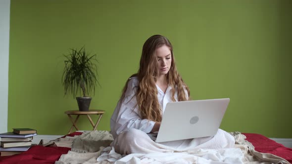 Young Girl Uses Laptop While Sitting On Bed In Apartment. alt