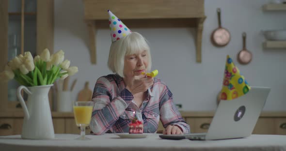 Cheerful Senior Woman Celebrates Birthday Online with Laptop, 70s Woman in Birthday Hat Blowing alt