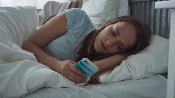 Tired Woman with Sad Eyes Using Smartphone on Bed at Home