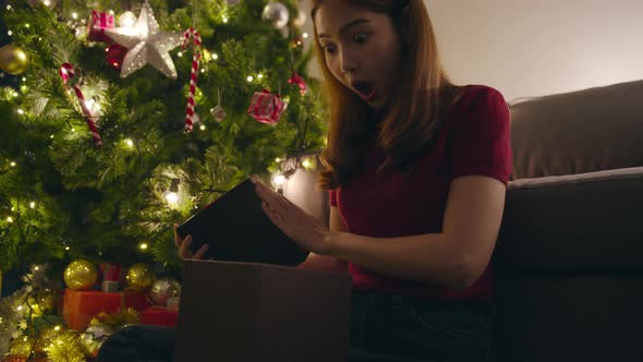 Young Asia female having fun opening X'Mas present box near Christmas tree decorated. alt